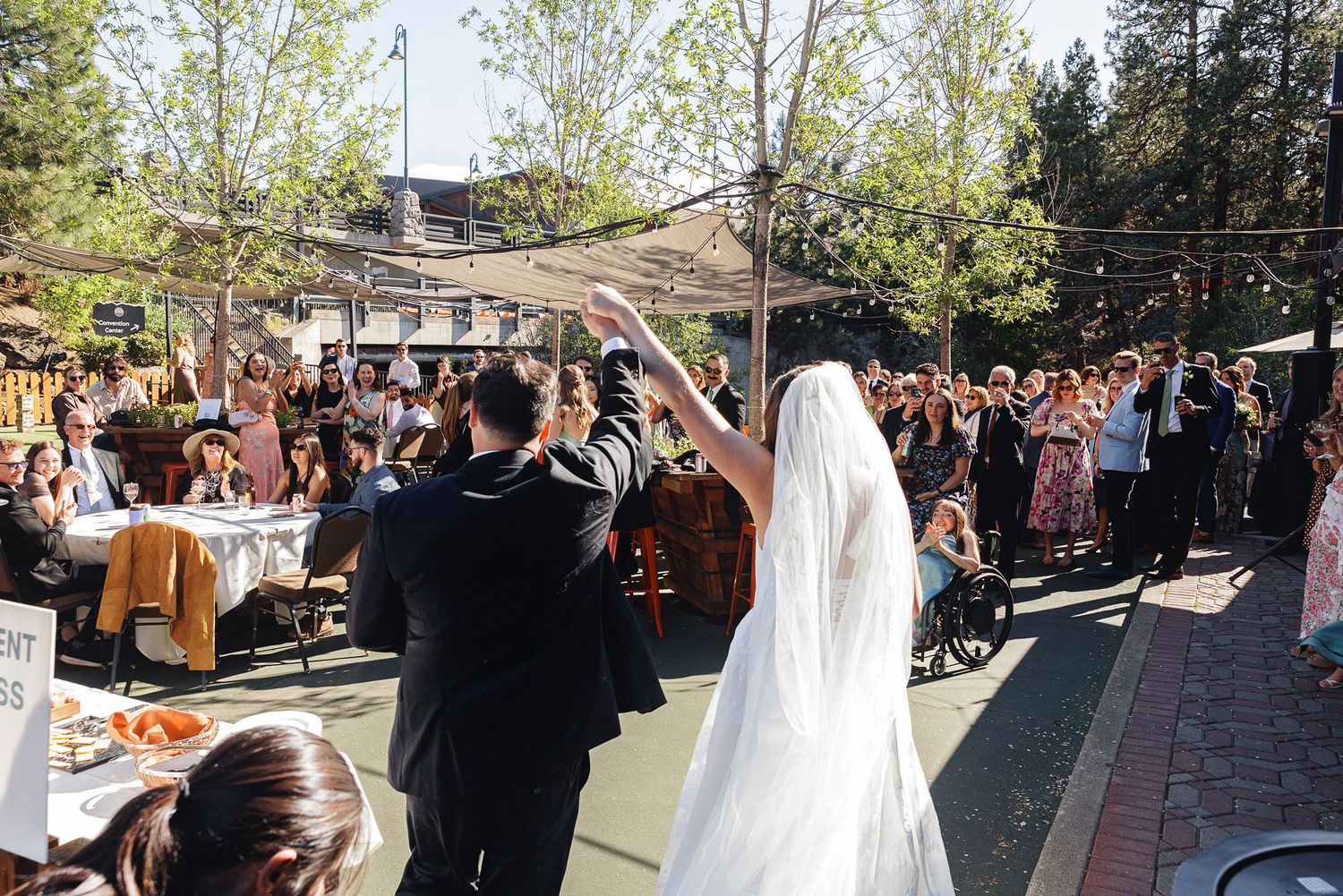 Bride and groom raise hands at outdoor wedding reception surrounded by cheering guests on sunny day.