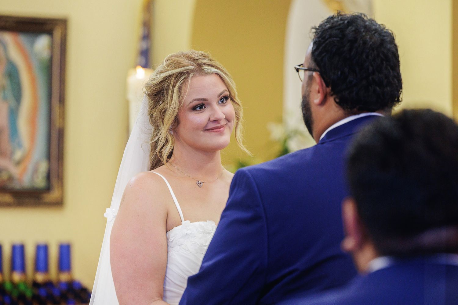 Bride in white dress and veil smiling at groom in blue suit during wedding ceremony in church.
