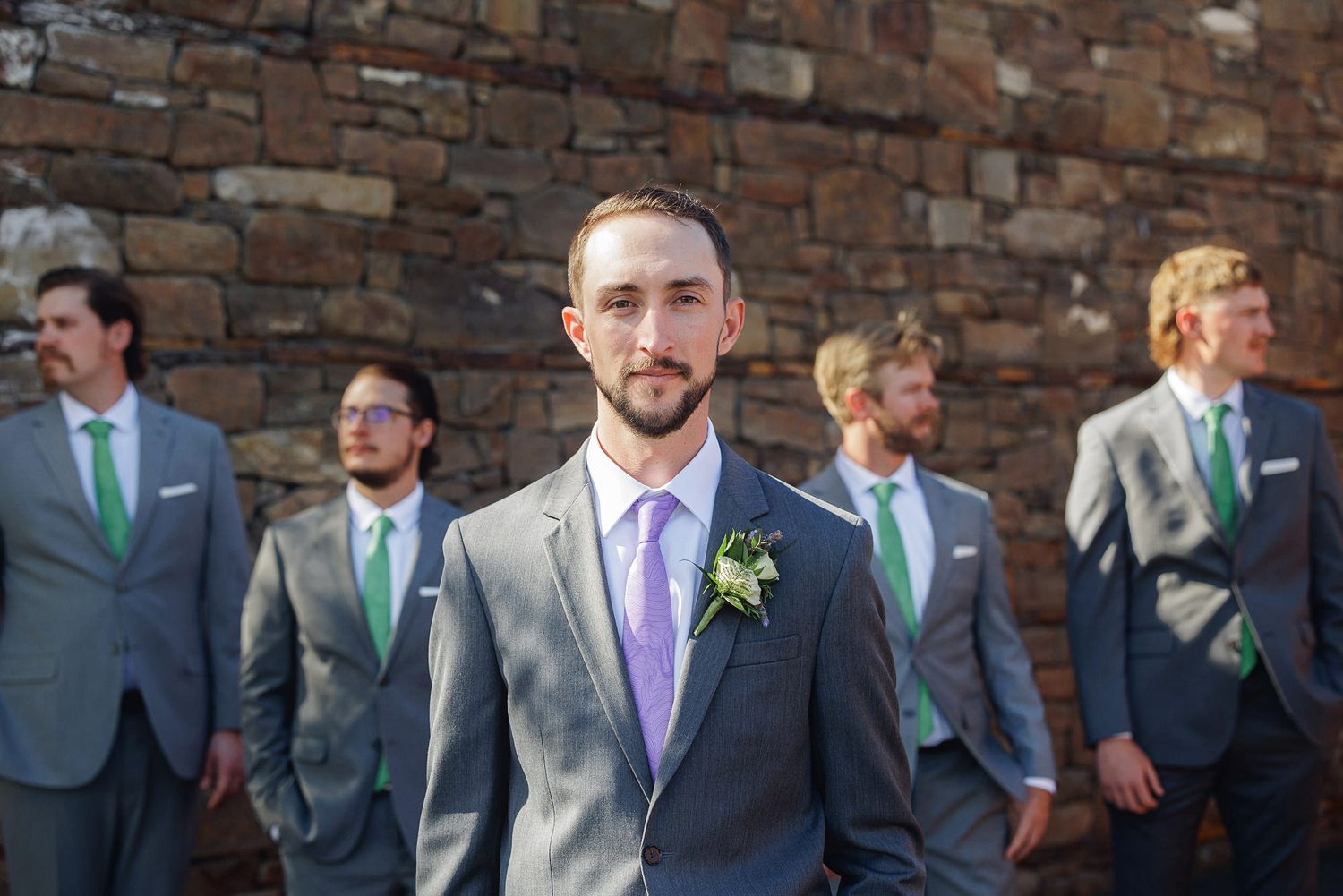 Groom in gray suit with purple tie and boutonniere stands before groomsmen in matching gray suits with green ties.