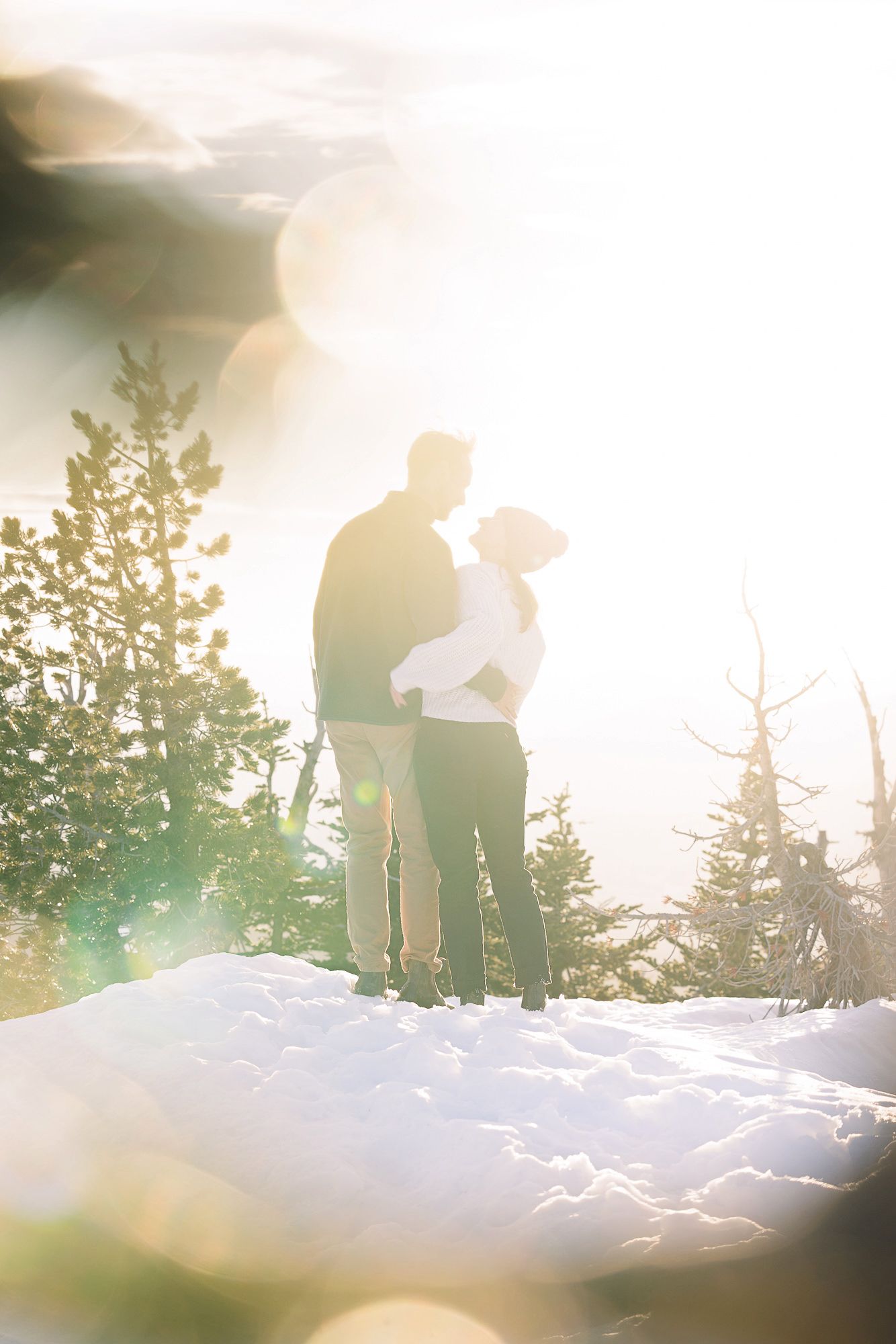 Couple embracing on snowy mountain summit surrounded by pine trees with bright golden sunlight flare in winter landscape.