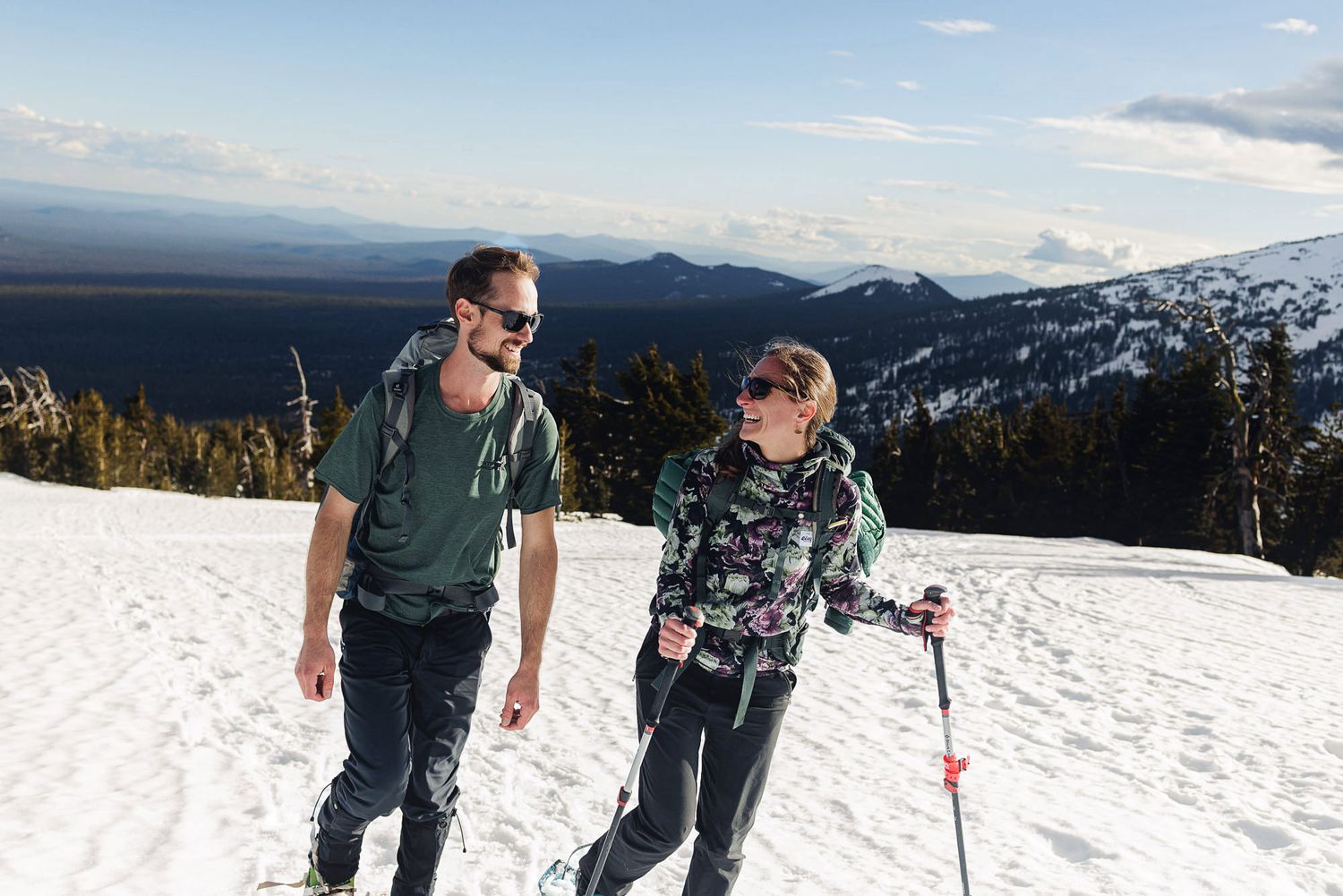 Two hikers walking on a snowy mountain trail with scenic views of forests and mountains in the background.