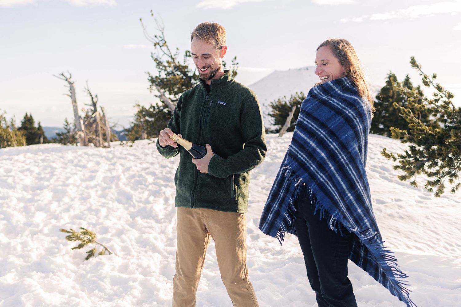 Couple standing in snowy mountain landscape, man in green jacket, woman wrapped in blue plaid blanket.