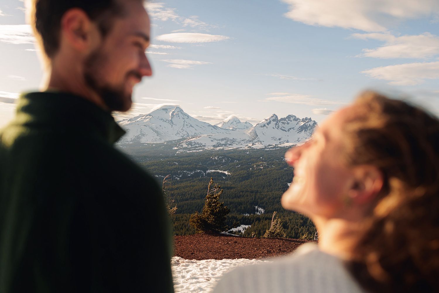 Couple laughing together outdoors with snow-capped mountain peaks and blue sky in the background.