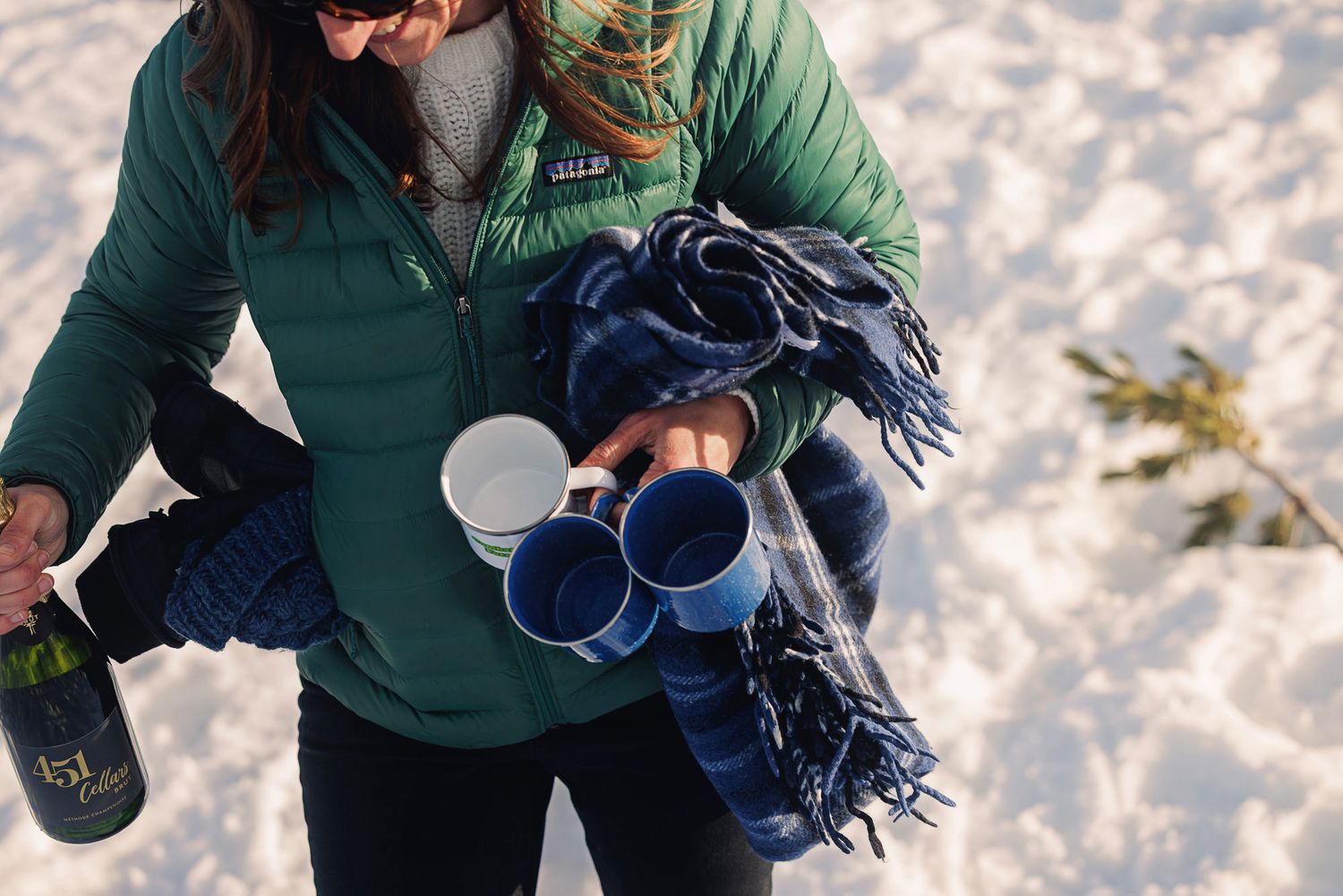 Woman in green jacket holding champagne bottle, blue mugs, and plaid blanket in snowy winter outdoor setting.