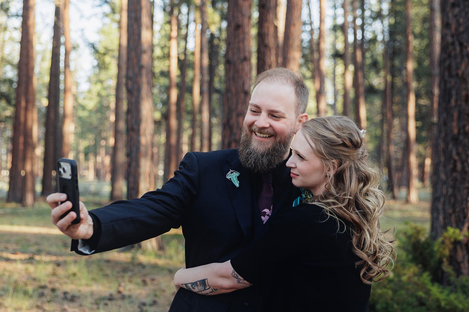 Couple taking a selfie in a pine forest, groom in black suit with floral pin, bride with curled hair.
