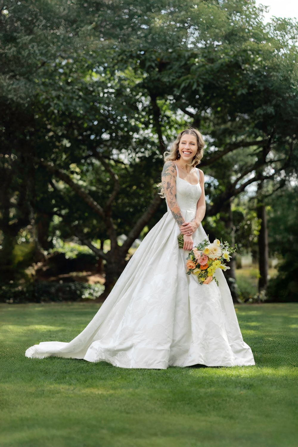 A wedding dress with elegant train and colorful bouquet photographed outdoors on a lush green lawn with trees in background.