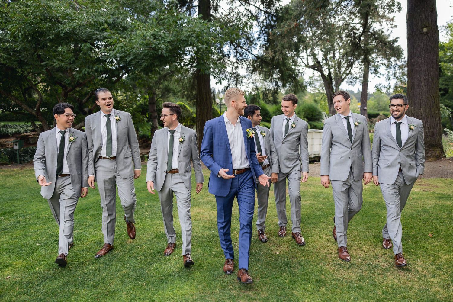 Groomsmen in coordinated grey suits walking together outdoors, with one member wearing a contrasting blue suit.