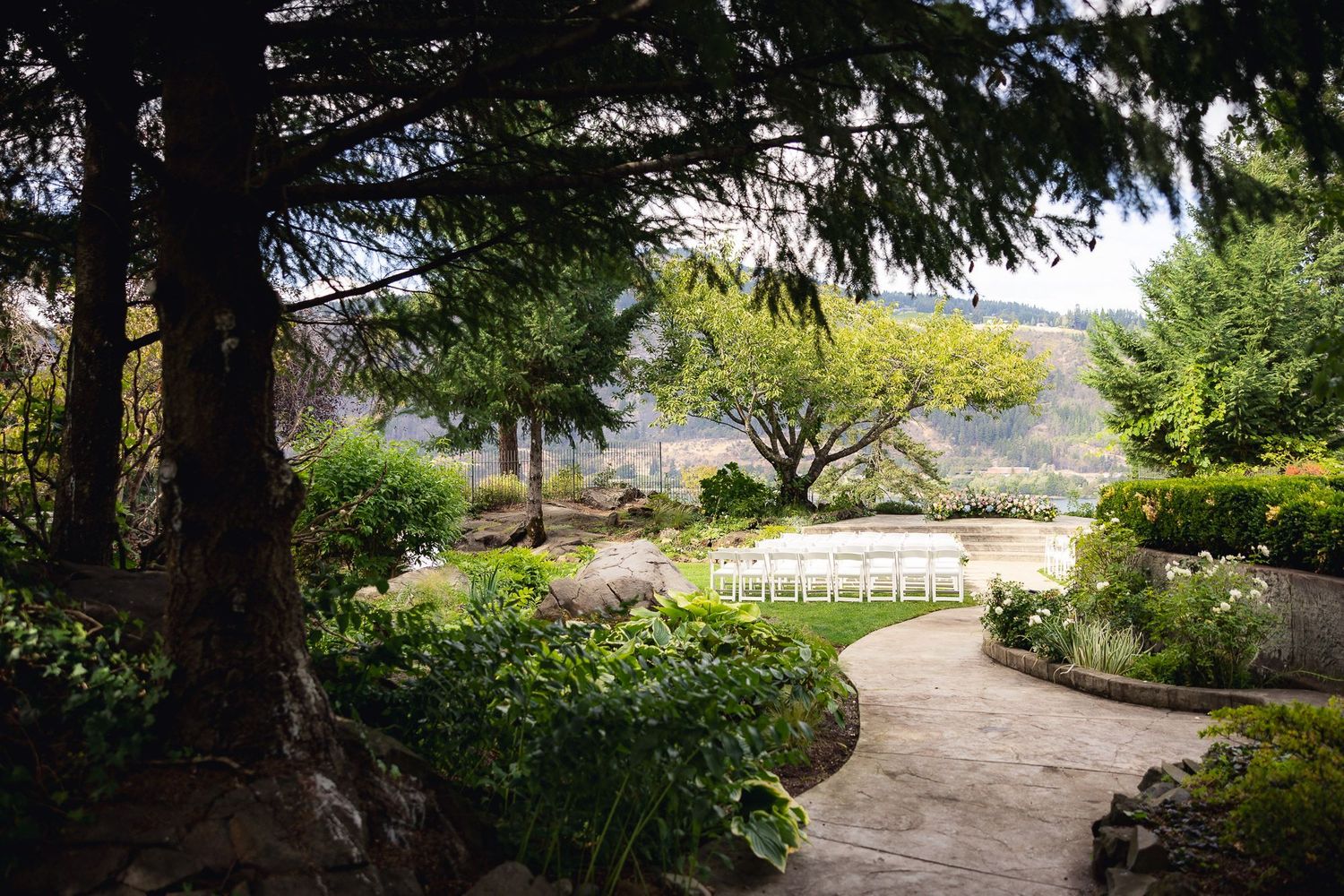 A winding garden path leads to an outdoor wedding ceremony setup with white chairs and mountain views in the distance.