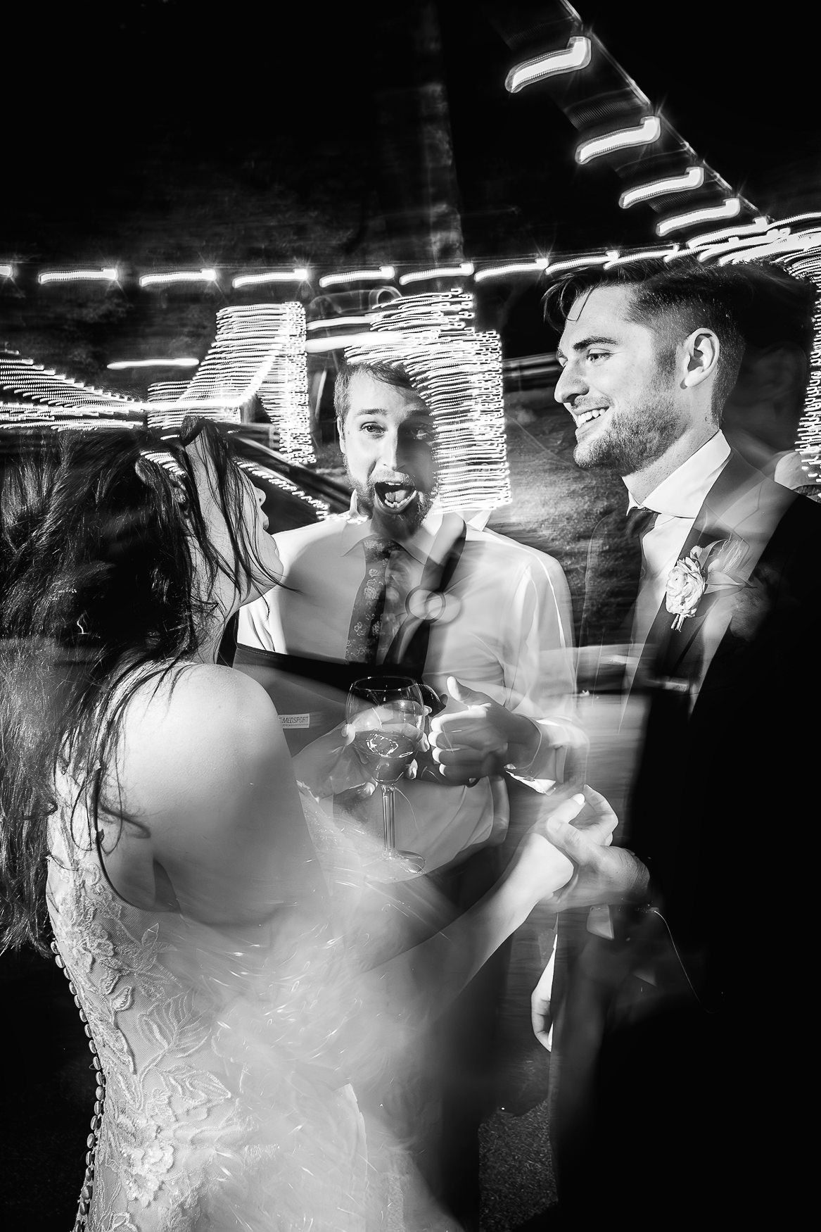 Black and white wedding photo showing celebratory dancing at a reception under string lights at night.