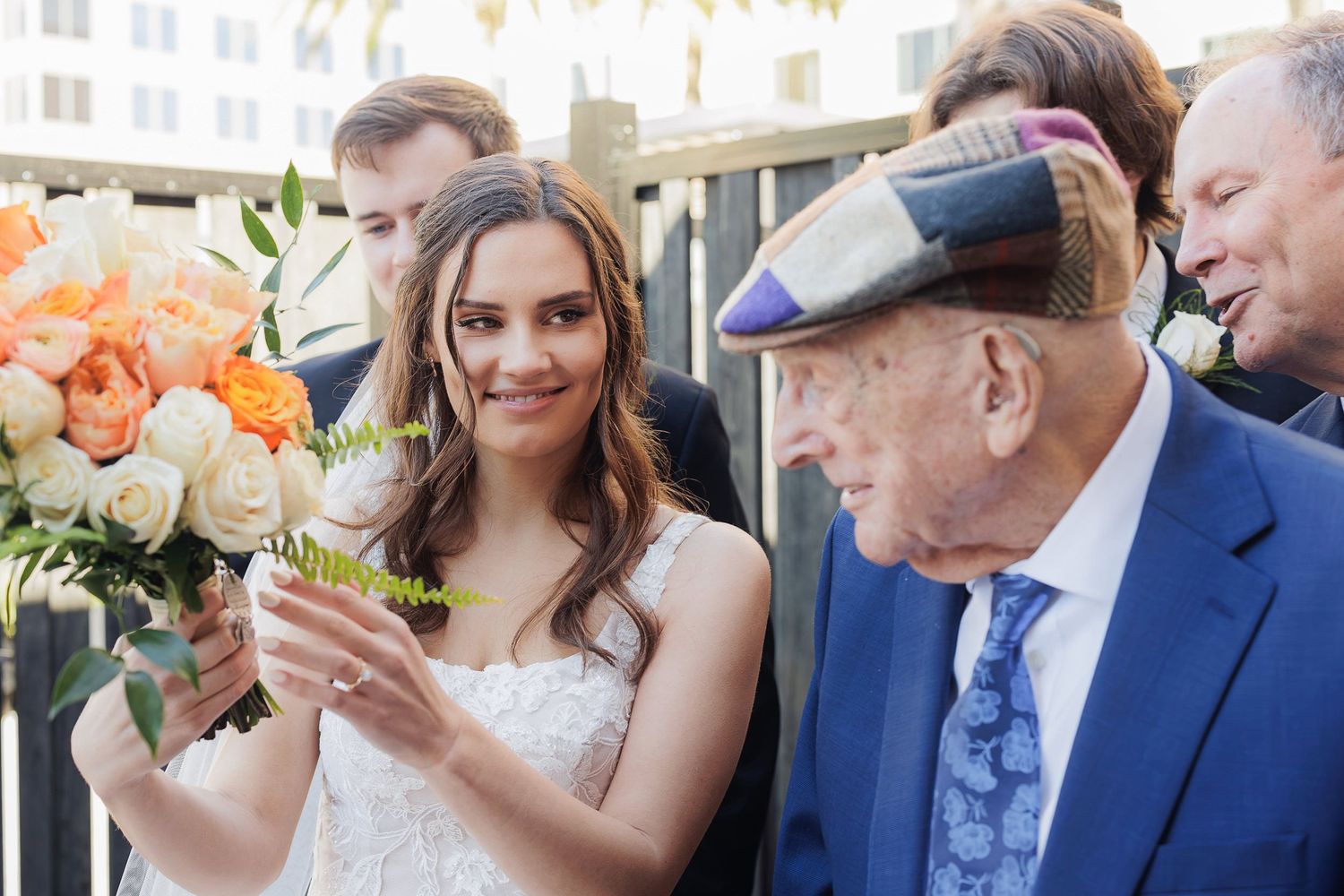 A bride in a white dress holds a bouquet while chatting with an elderly gentleman in a blue suit and cap at a wedding.
