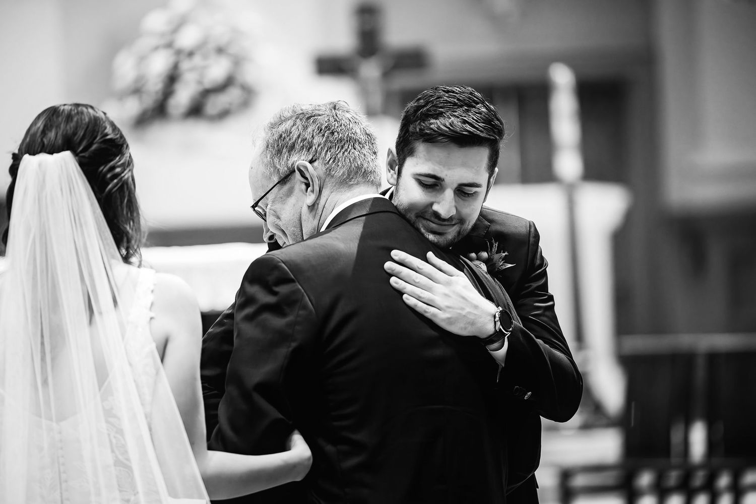 An emotional black and white moment at a wedding ceremony in a church shows the groom embracing a figure.