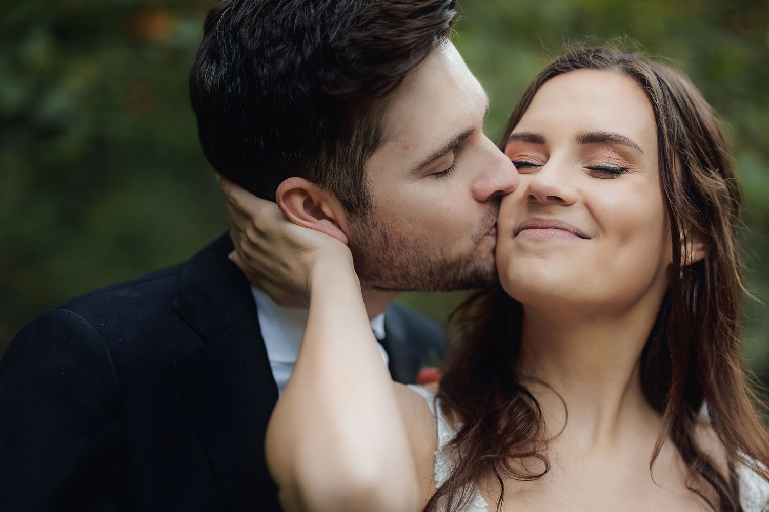 A romantic moment between a couple sharing a tender kiss in an outdoor setting with lush greenery in the background.