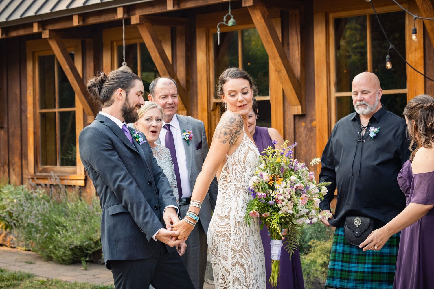 A bride holding a colorful wildflower bouquet laughs joyfully during an outdoor wedding ceremony at a rustic venue.
