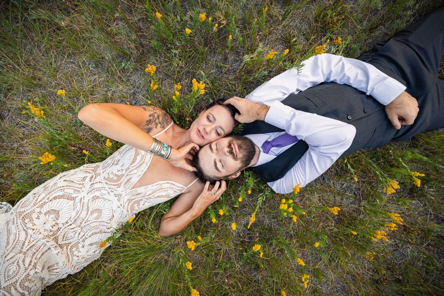 A couple in wedding attire lies on grass surrounded by yellow wildflowers in an overhead view.