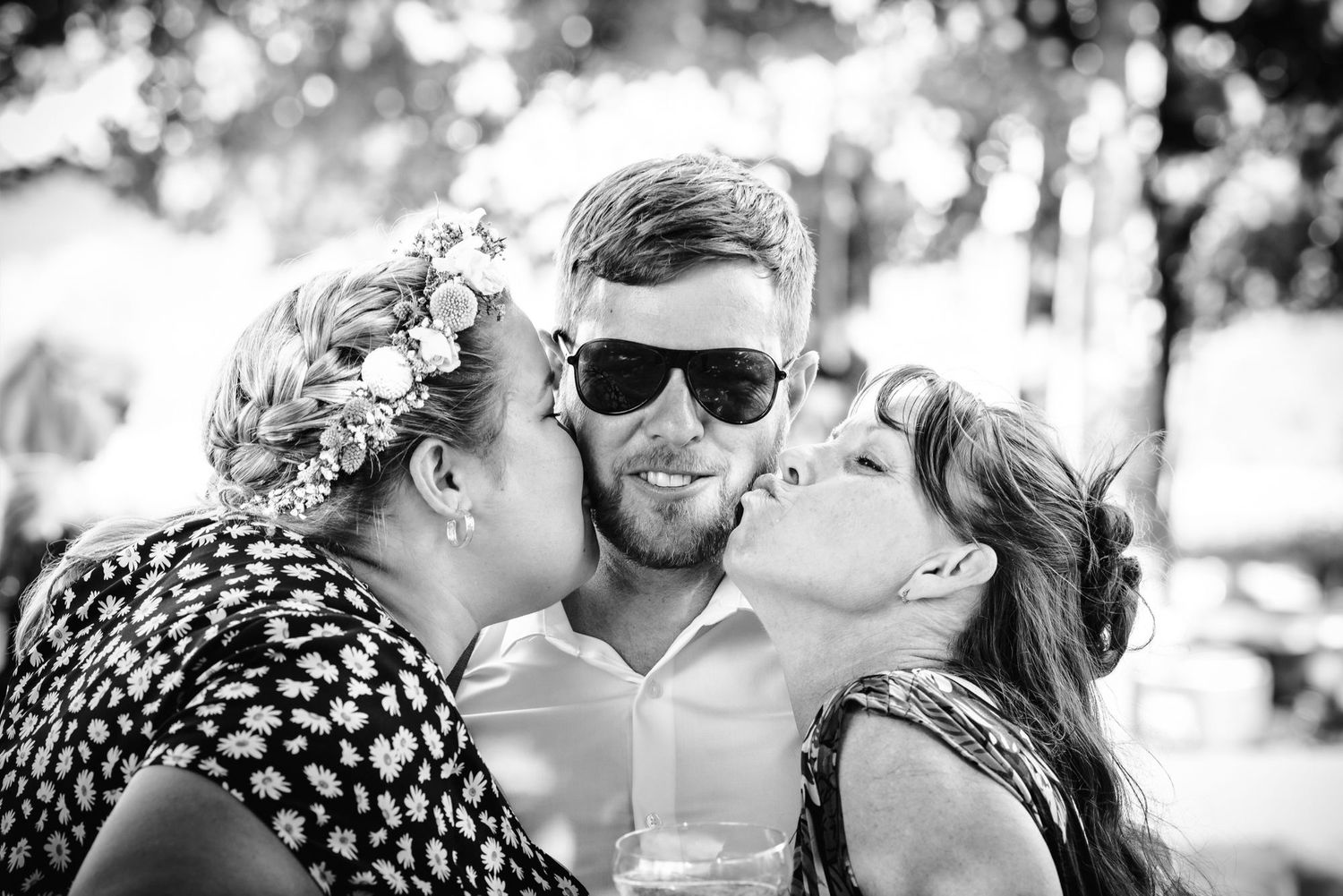 A candid black and white photo captures a fun moment between three people sharing kisses and laughter outdoors.