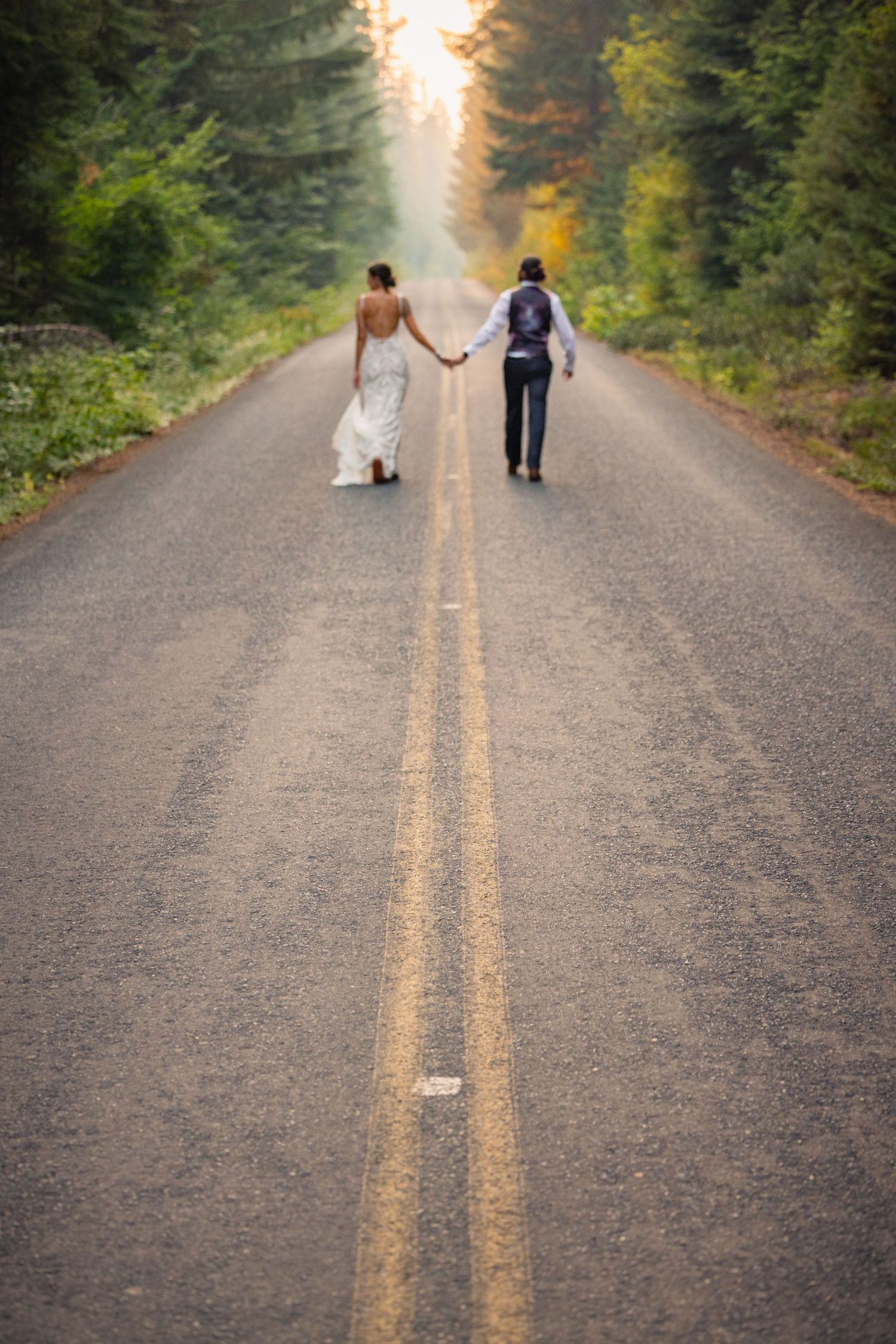 Two newlyweds walk hand in hand down a forest road illuminated by golden sunlight.