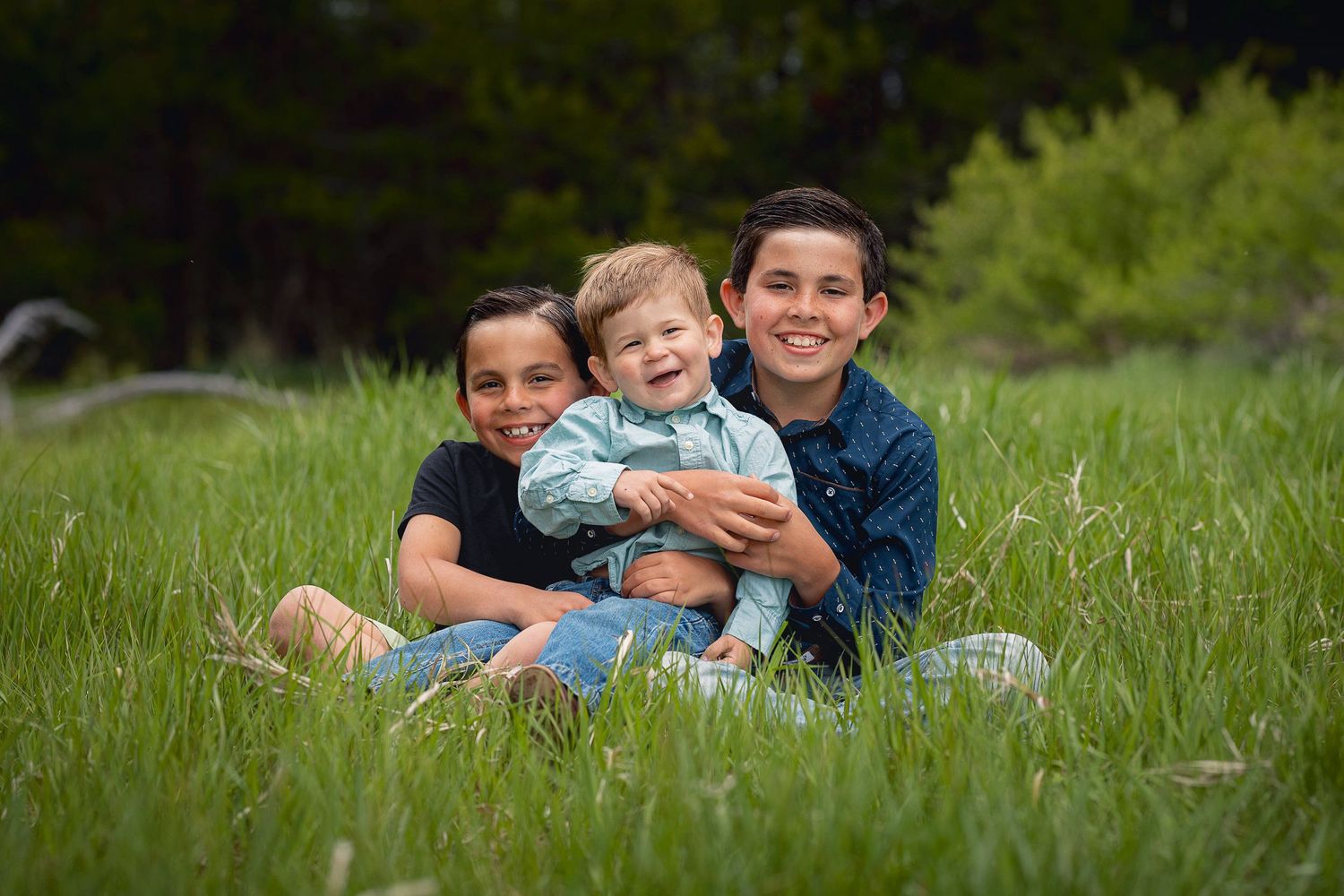 Three people sitting close together and smiling in a lush green field on a sunny day.