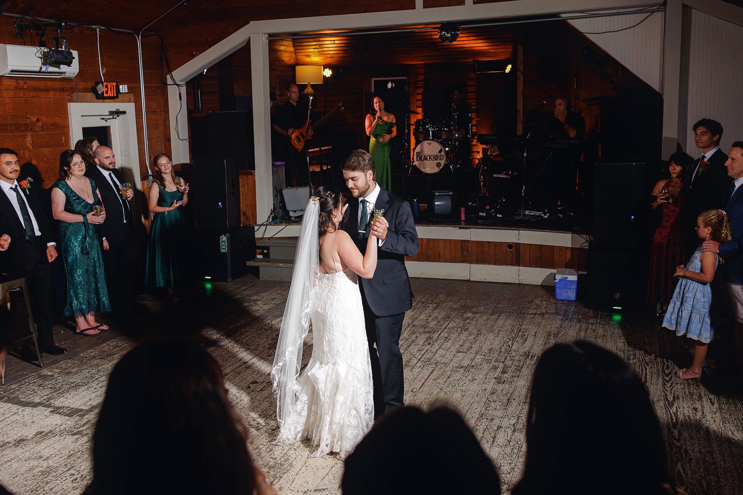 Bride and groom share their first dance at wedding reception while guests watch and live band performs on stage.
