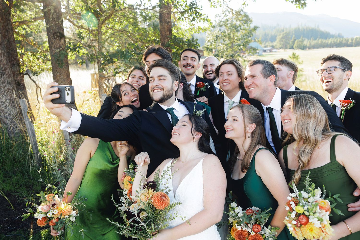 Wedding party taking a group selfie outdoors, bridesmaids in green dresses holding colorful bouquets with mountains behind.