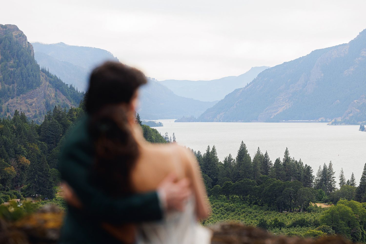A couple embraces overlooking a stunning mountain lake vista with lush evergreen forests in the Columbia River Gorge.
