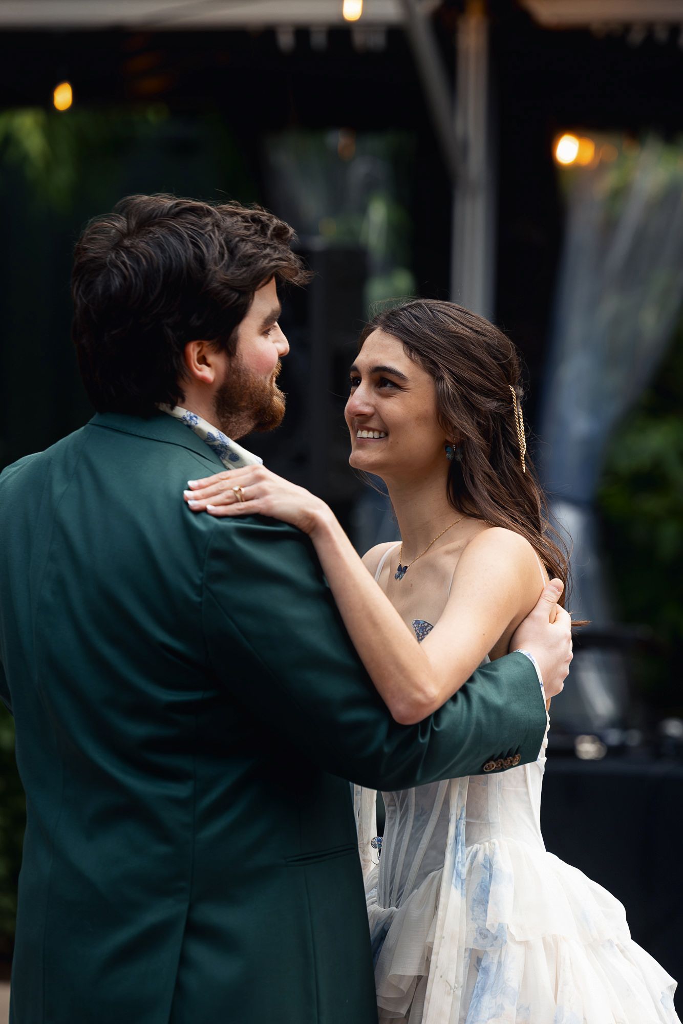 A smiling bride in a white gown shares a first dance with a groom in a green suit at an outdoor wedding reception.