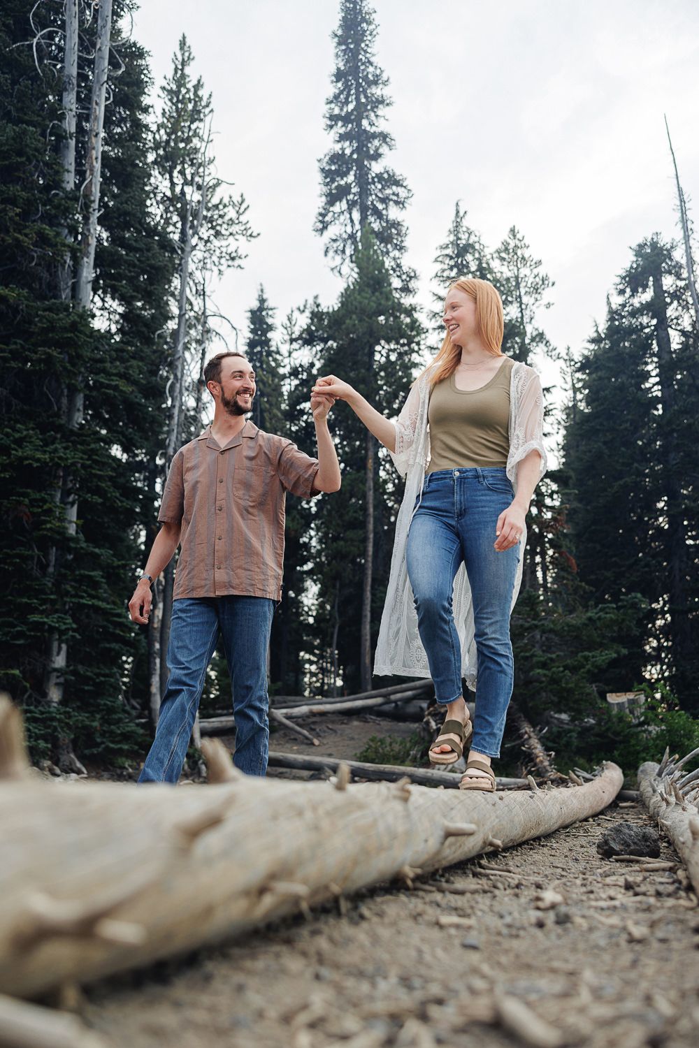 Two people walking and balancing along a fallen tree trunk in a forest with tall evergreen trees in the background.