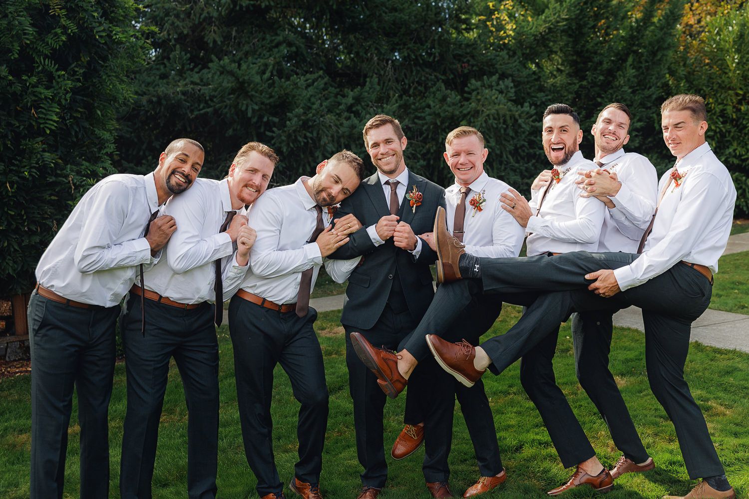 A group of groomsmen in white shirts and dark pants pose playfully together in an outdoor setting.