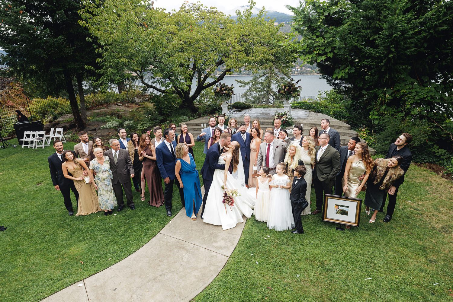 Large wedding party poses outdoors on green lawn with scenic waterfront and lush trees in background.