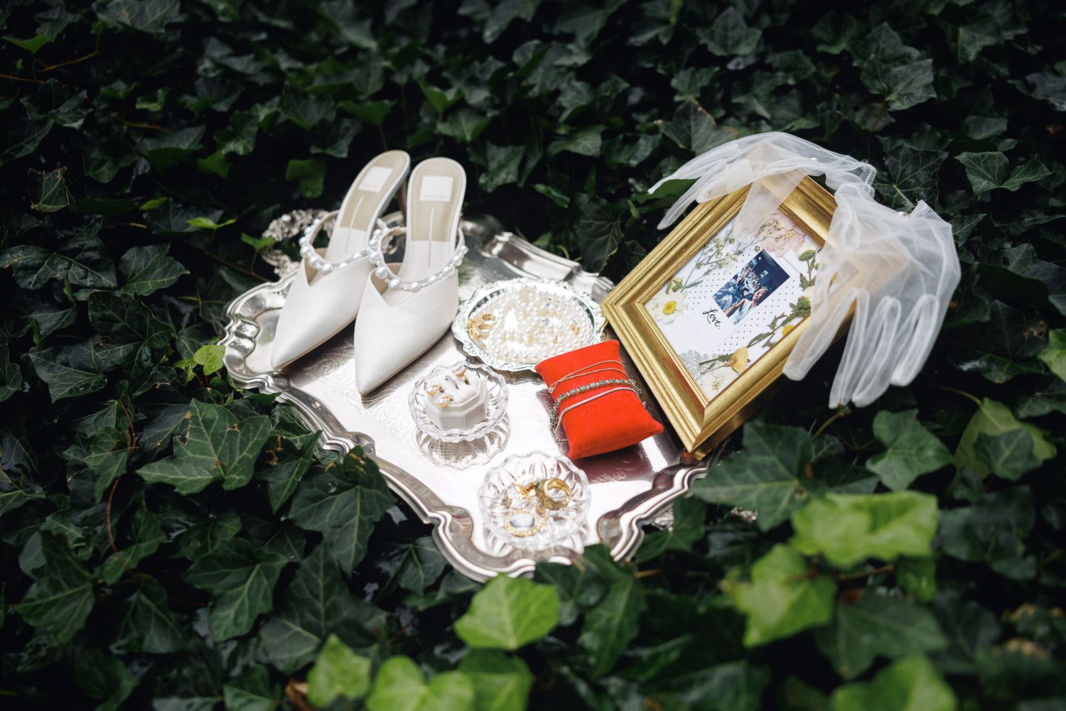 Silver tray with bridal accessories including white heels, gloves, red ring box, and framed photo on ivy background.