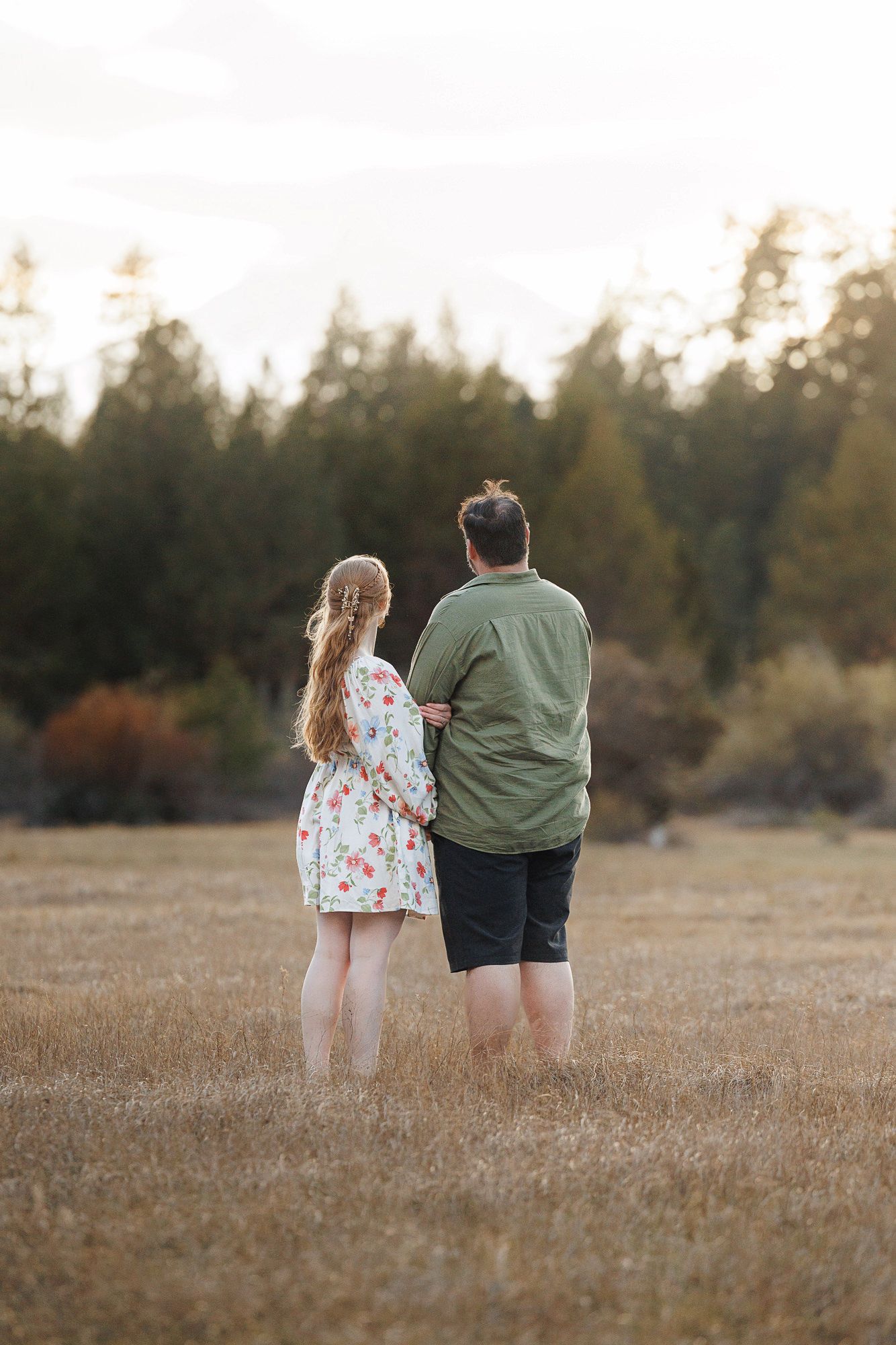 Couple standing in a golden field facing pine trees, woman in floral dress and man in green shirt with arms around each other.