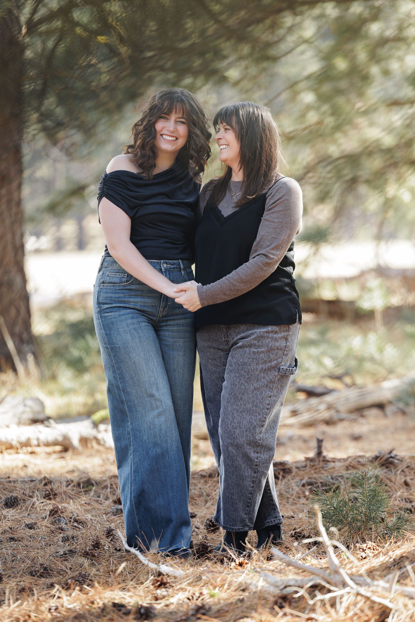 Two smiling women with dark hair embrace outdoors among pine trees, wearing casual black and gray outfits.