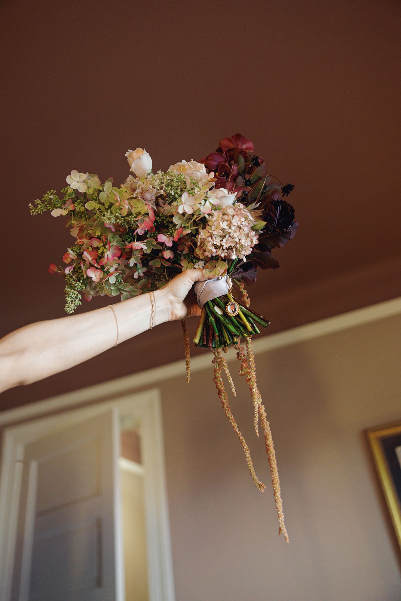 A hand holds a lush bridal bouquet with burgundy, blush, and green blooms with trailing amaranthus accents.