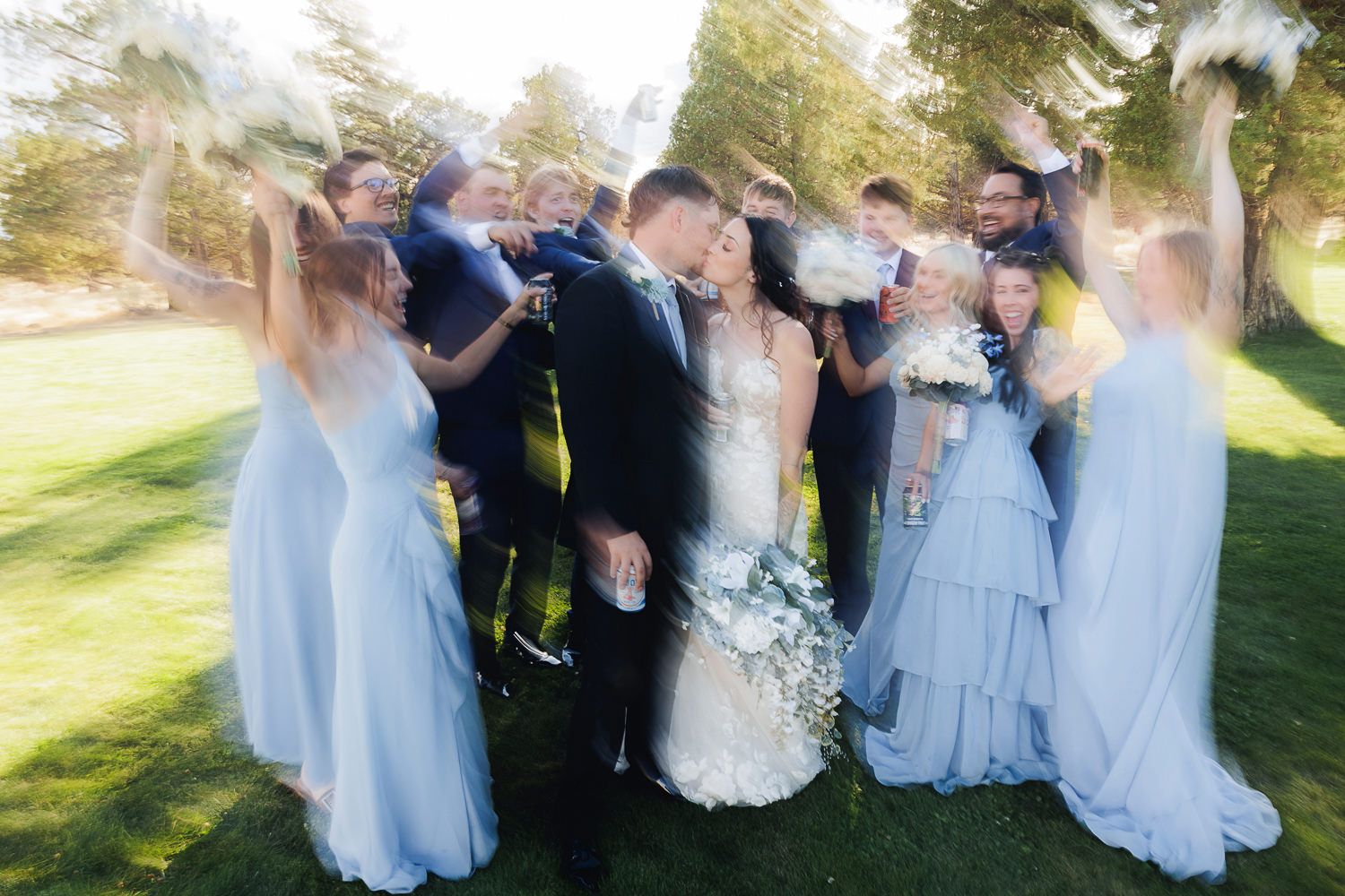 A wedding party celebrates and laughs together on a sunny lawn in soft focus light blue dresses and formal attire.