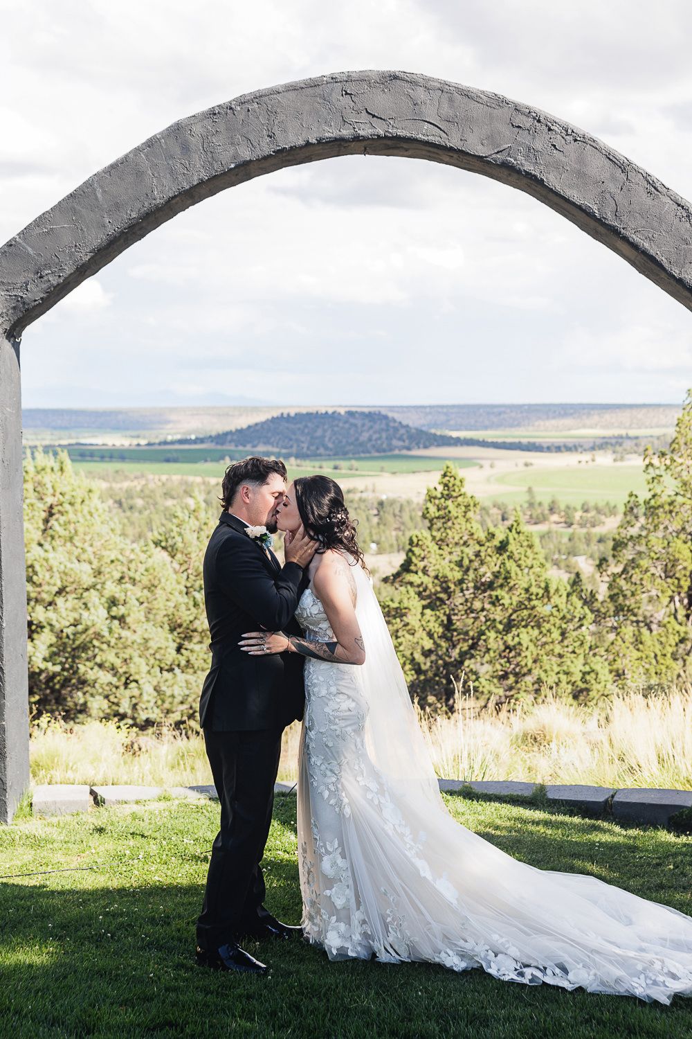 A couple shares an intimate wedding moment under a stone arch overlooking scenic mountain views and greenery.