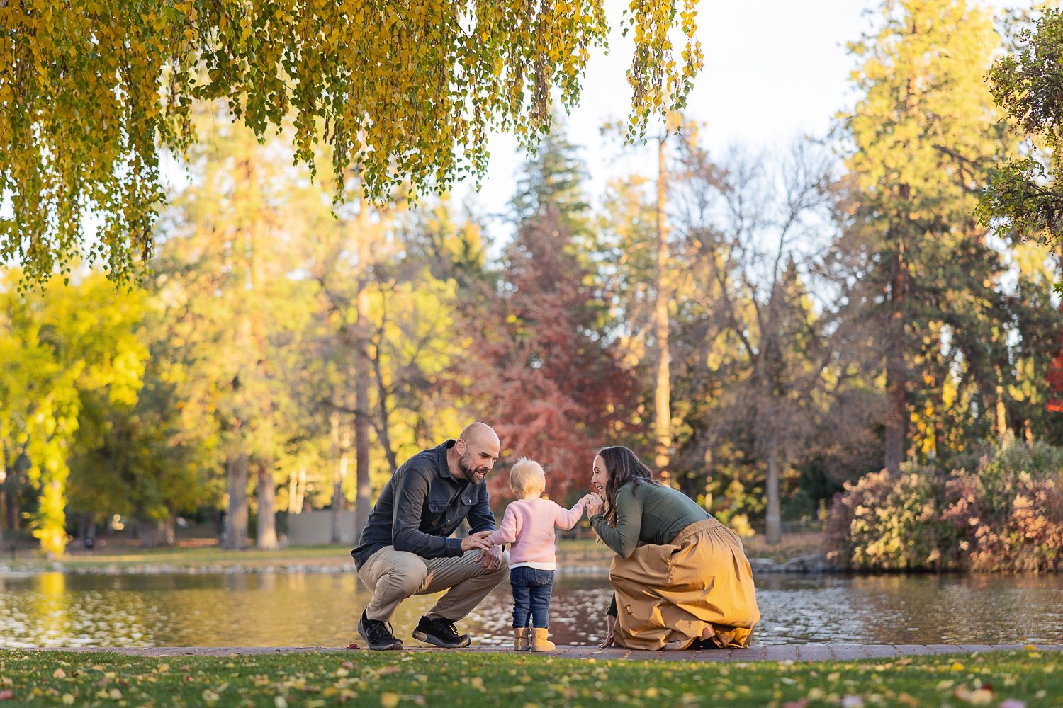Family enjoys autumn picnic by a pond surrounded by colorful fall foliage and golden weeping willow trees.