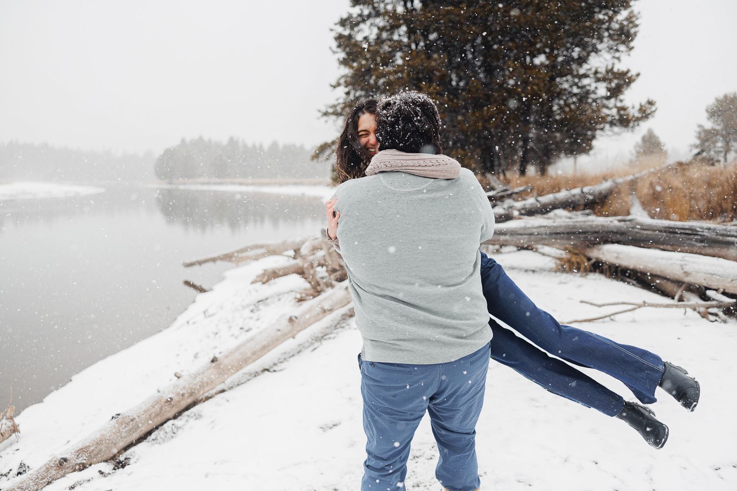 A couple embraces in a snowy winter landscape with a lake and trees in the background.