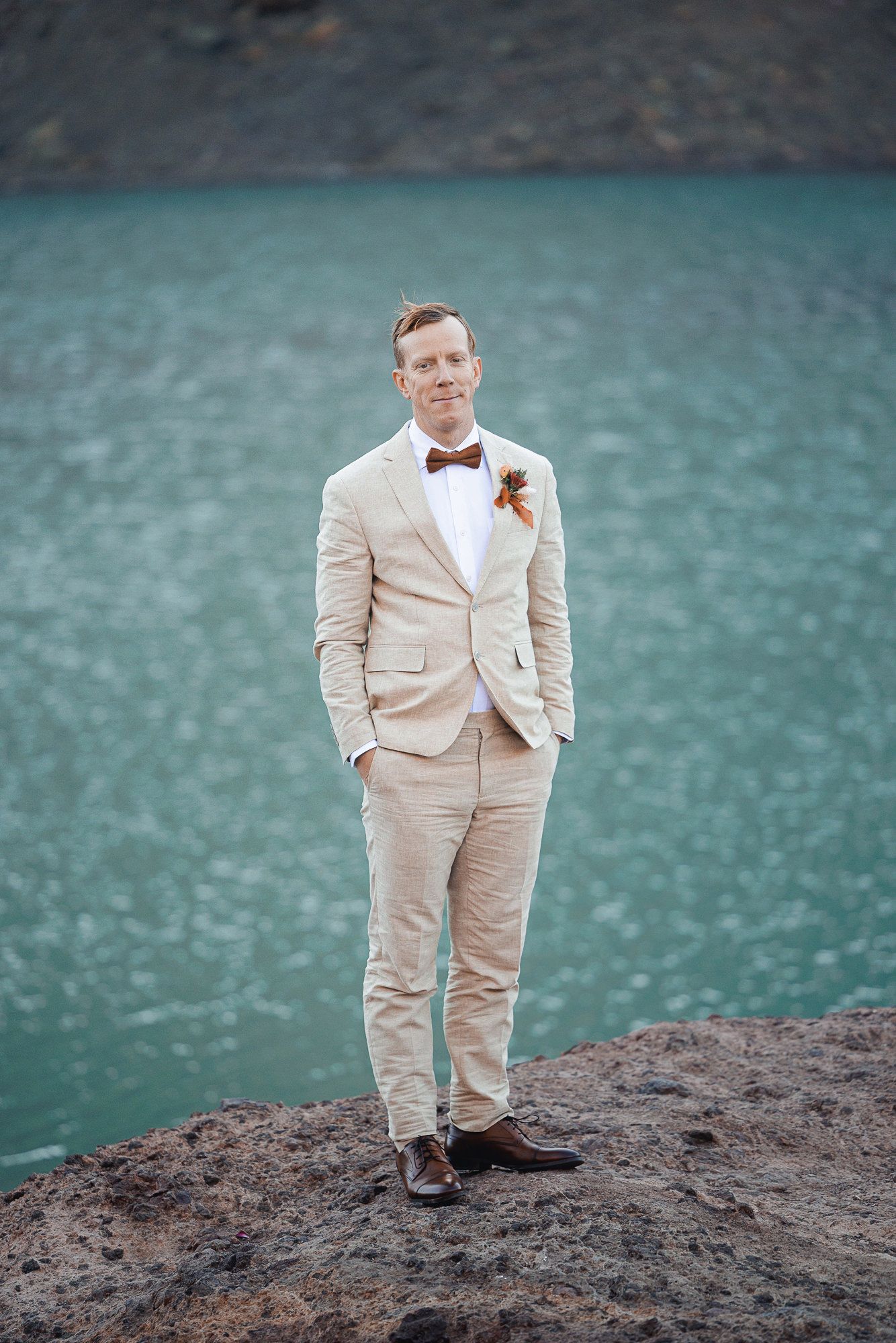 Groom in a tan linen suit, brown bow tie and floral boutonniere standing on a rocky ledge above a turquoise lake.