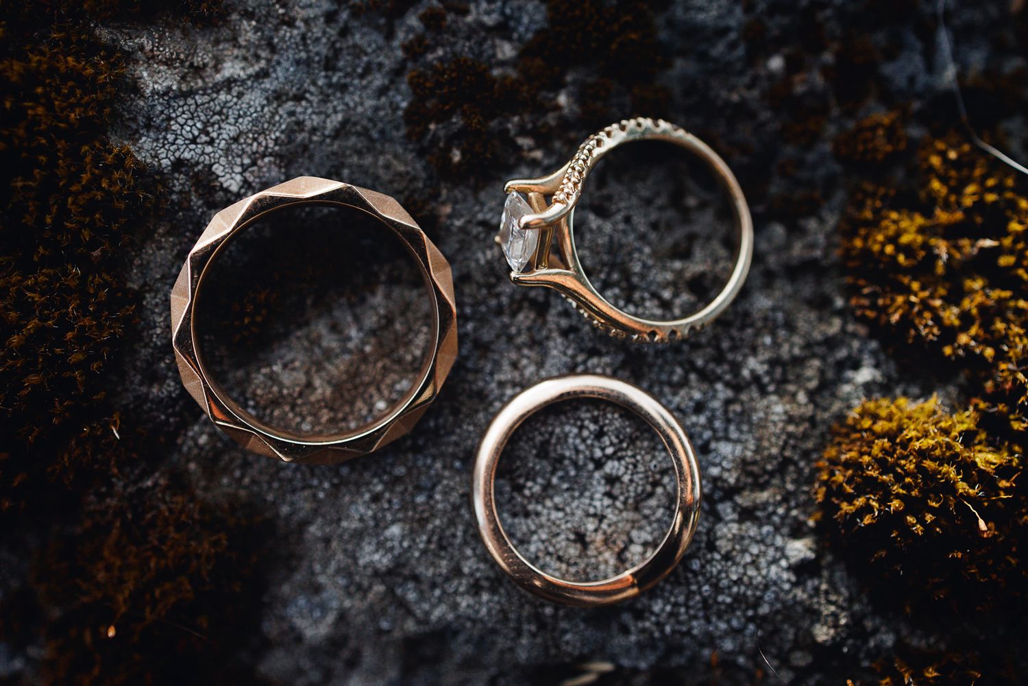 Three rose gold wedding rings displayed on a mossy rock surface, including a diamond engagement ring.