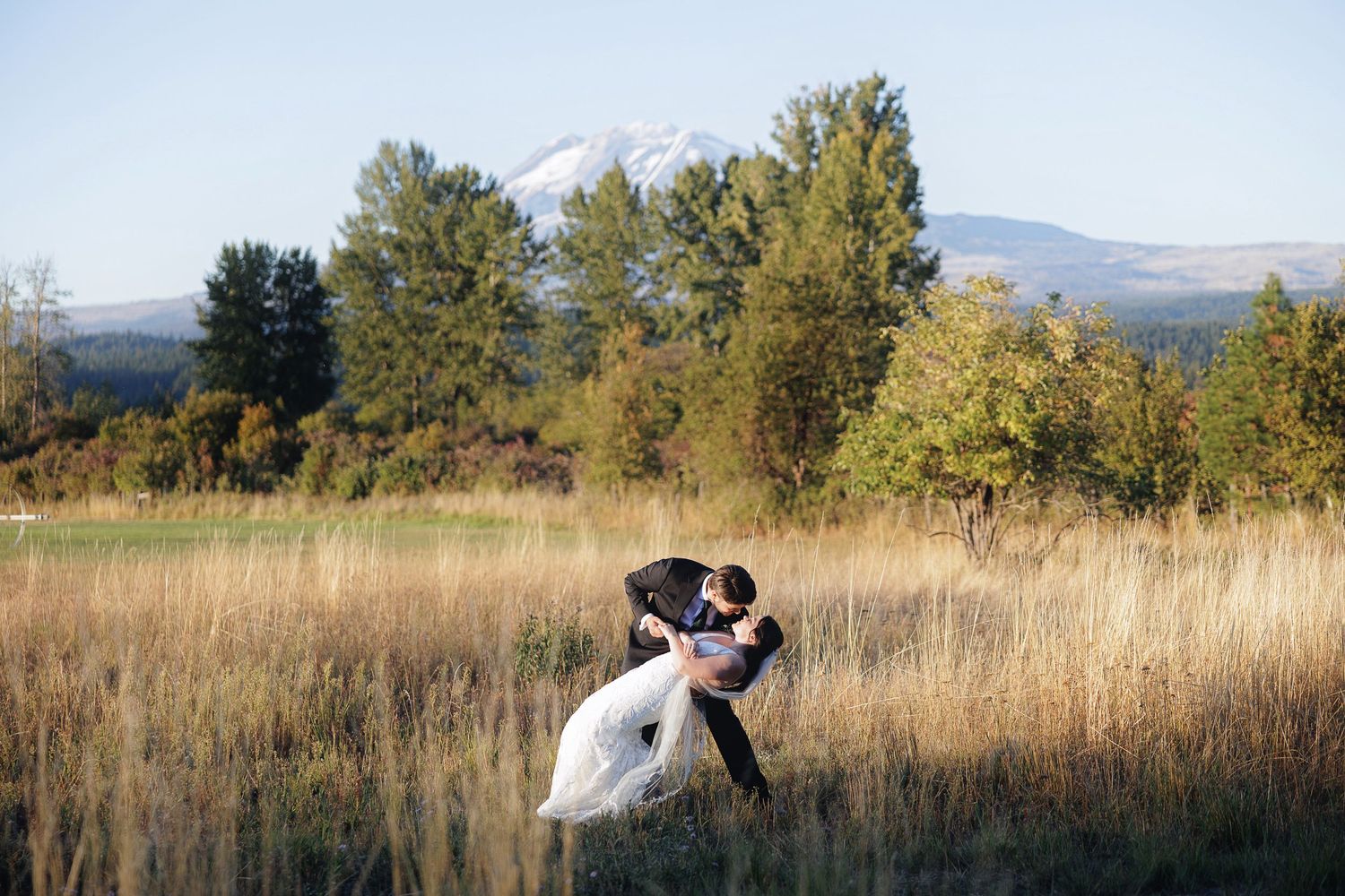 Groom dips bride in golden field with snow-capped mountain and evergreen trees in background at outdoor wedding.