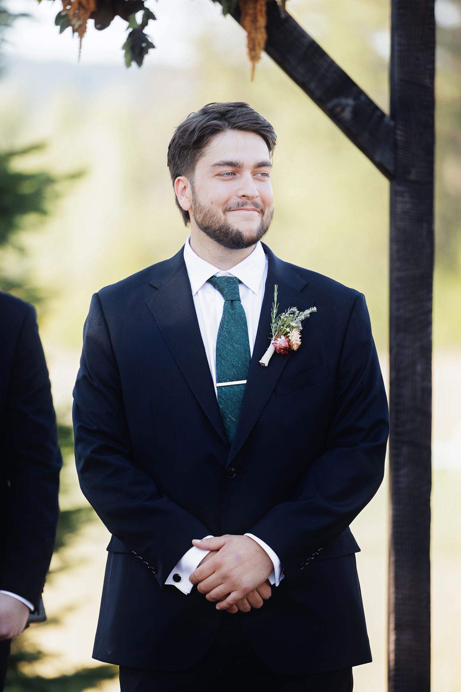 Groom in navy suit with green tie and boutonniere smiling at wedding ceremony outdoors.