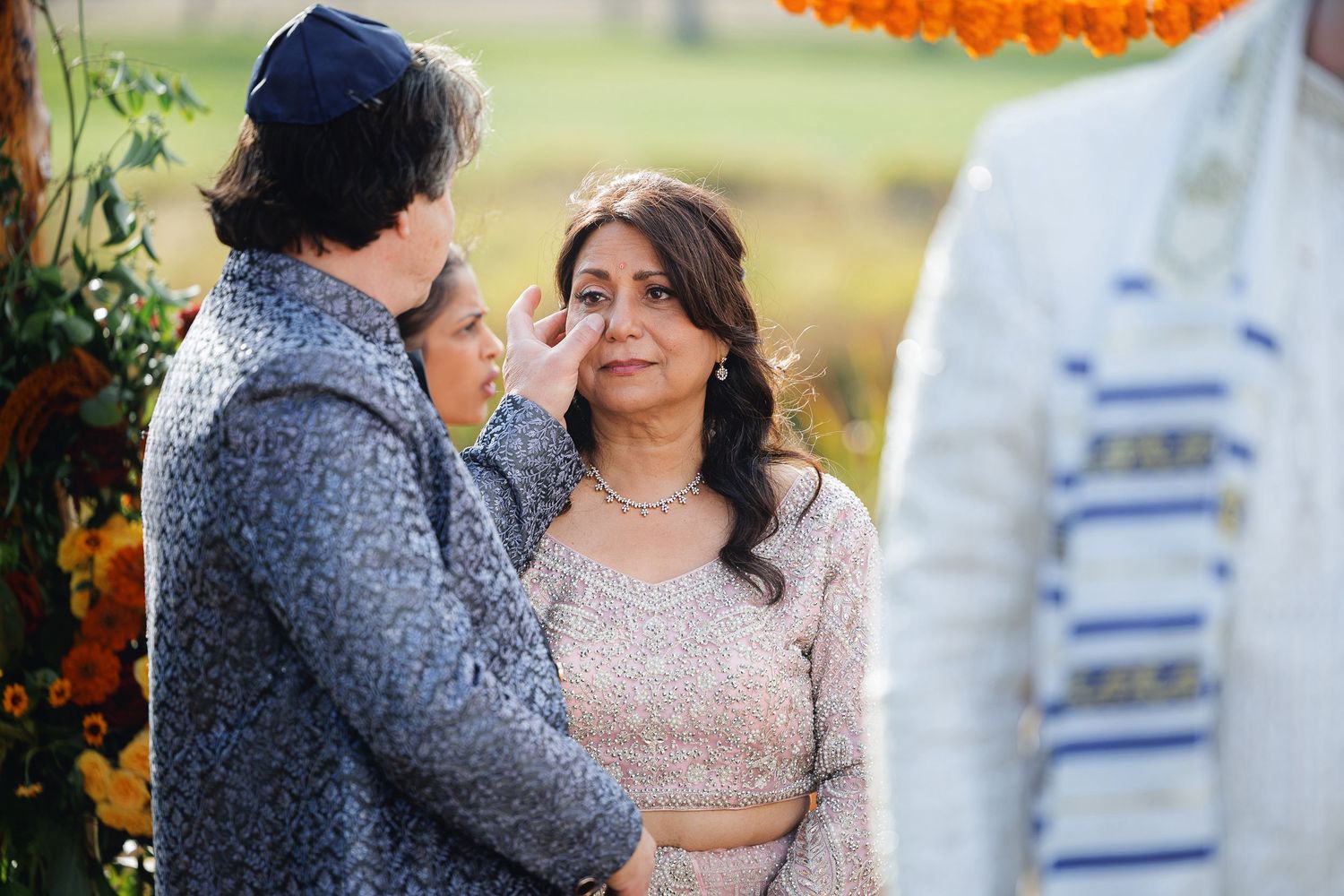 Groom gently touches bride's face during an outdoor South Asian wedding ceremony with marigold floral backdrop.