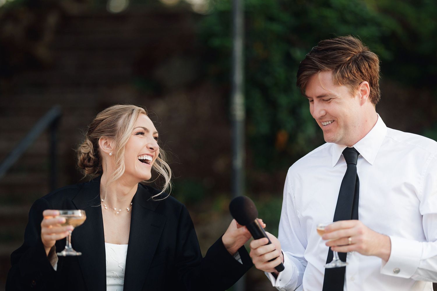 A laughing bride and groom in formal attire share a joyful moment holding champagne glasses at a wedding reception.