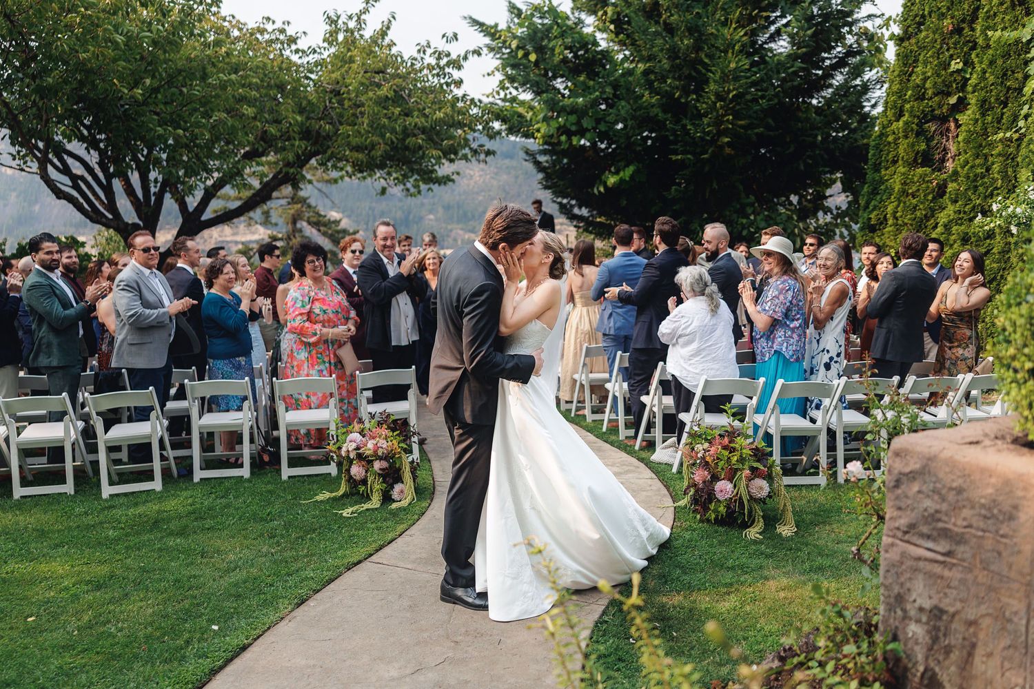 Bride and groom share a romantic kiss at outdoor wedding ceremony with guests seated in white chairs on lush green lawn.