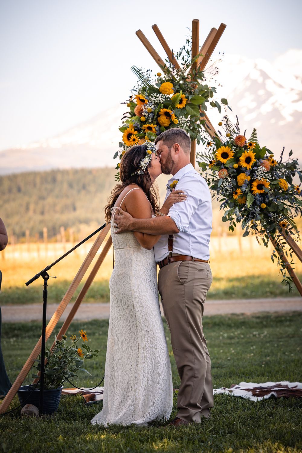 A couple embraces under a wooden arch decorated with sunflowers and greenery at an outdoor rustic mountain wedding ceremony.