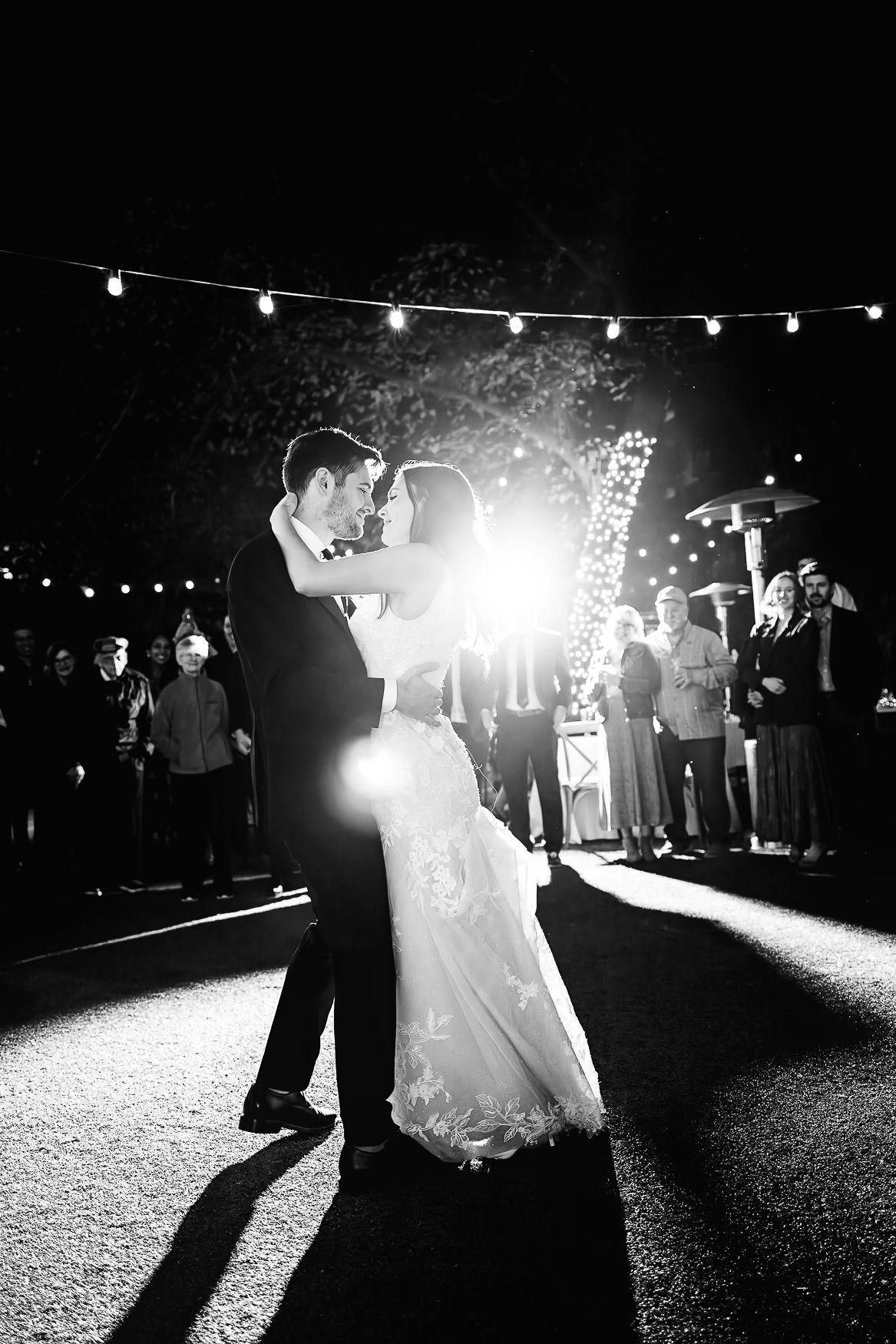 A romantic wedding couple shares their first dance under twinkling lights at an outdoor evening reception.