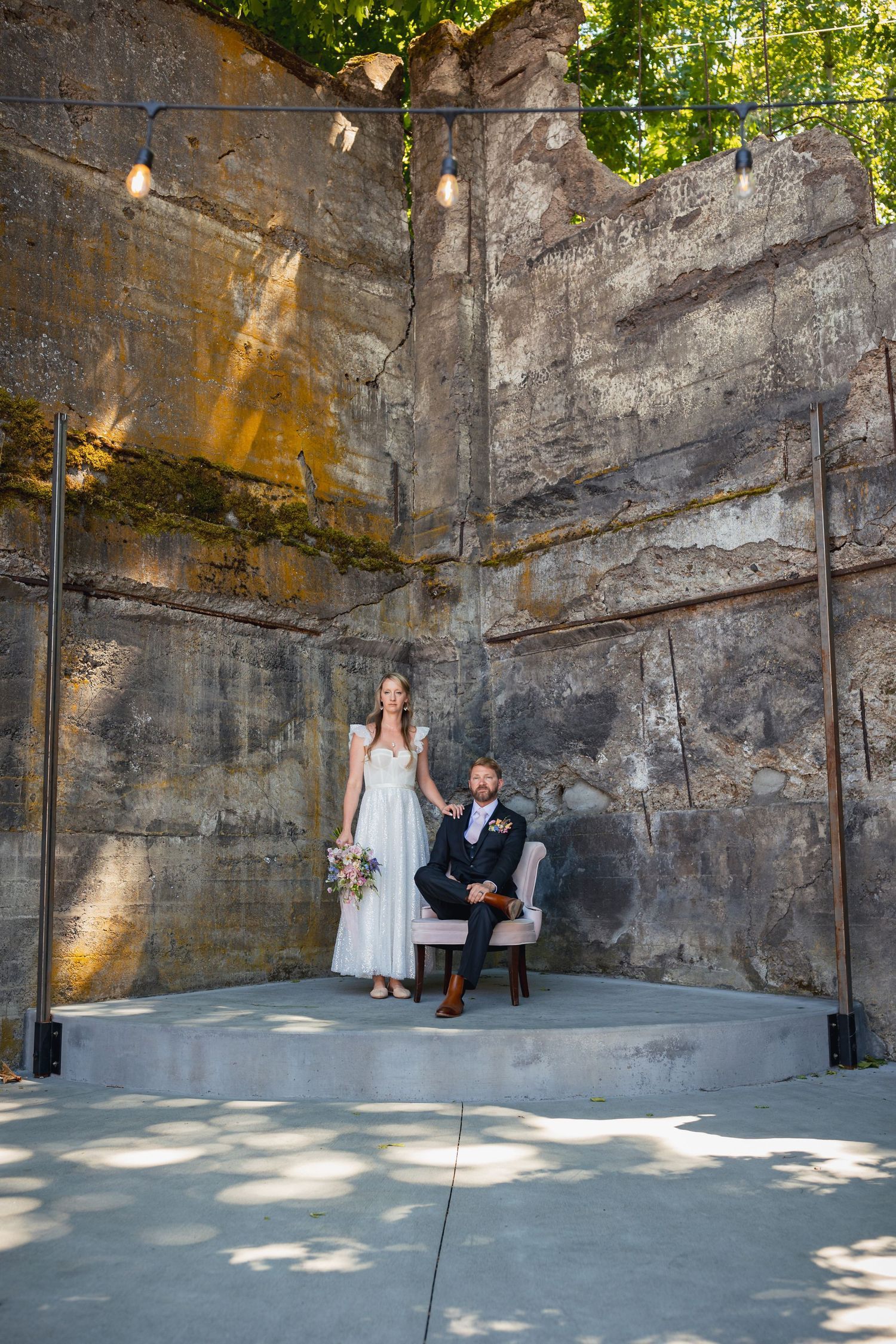 A couple poses for a wedding photo against a tall stone wall in an outdoor setting with dappled sunlight.