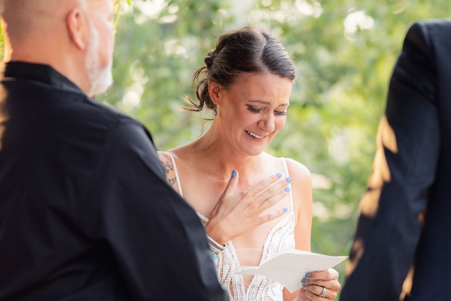 A bride in a white lace dress reads from a paper during an outdoor wedding ceremony.