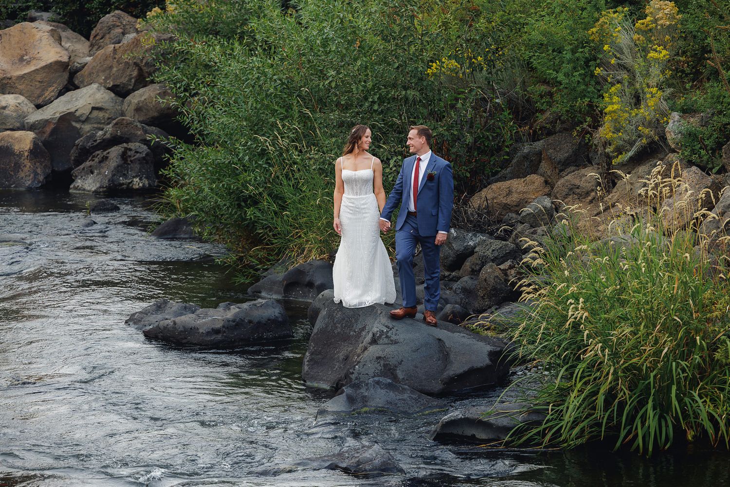 Wedding couple stands on rocky riverbank surrounded by lush greenery and flowing water in natural outdoor setting.