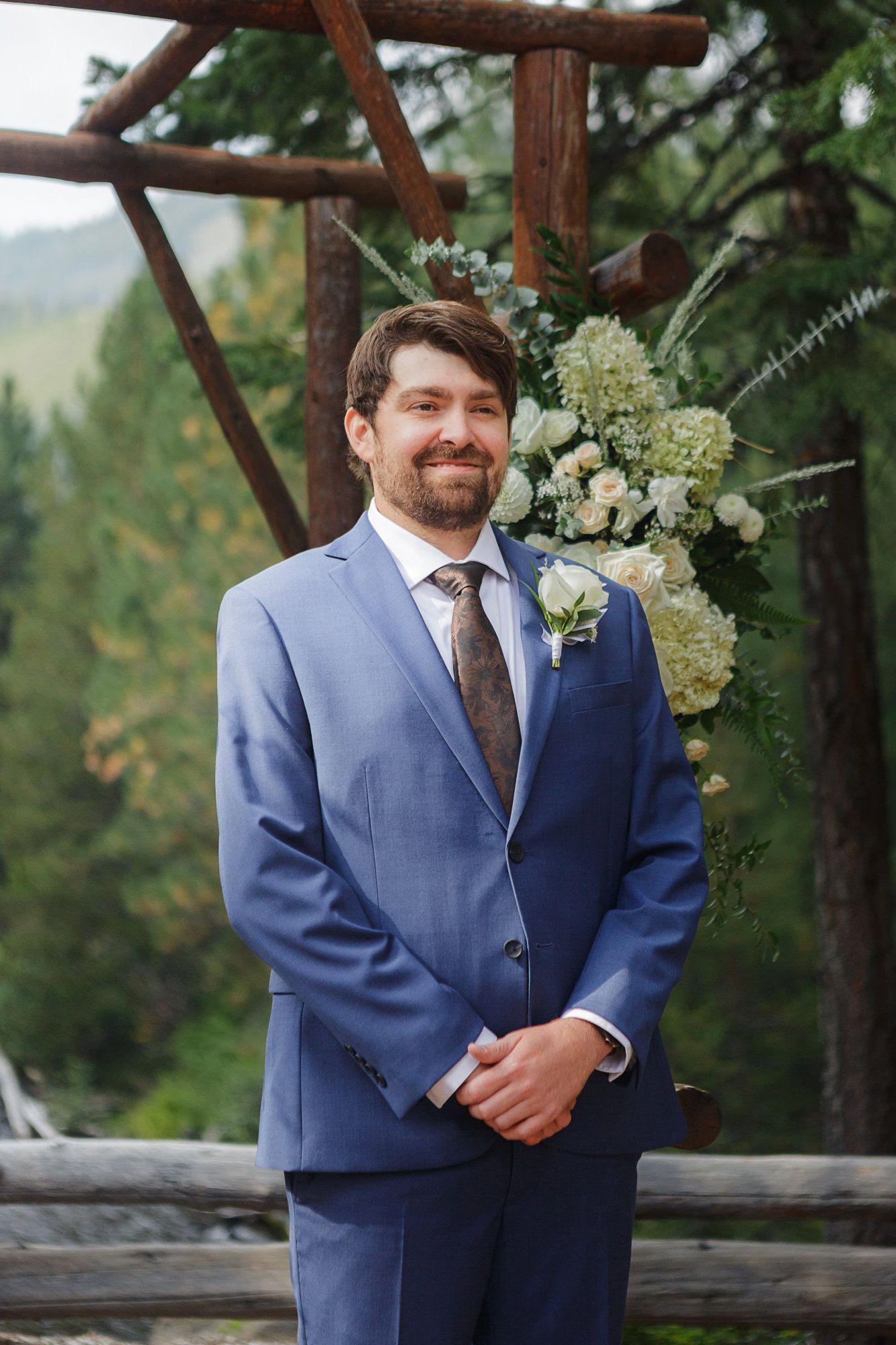 A groom in a blue suit and brown tie stands at a rustic wooden railing with white floral decorations.