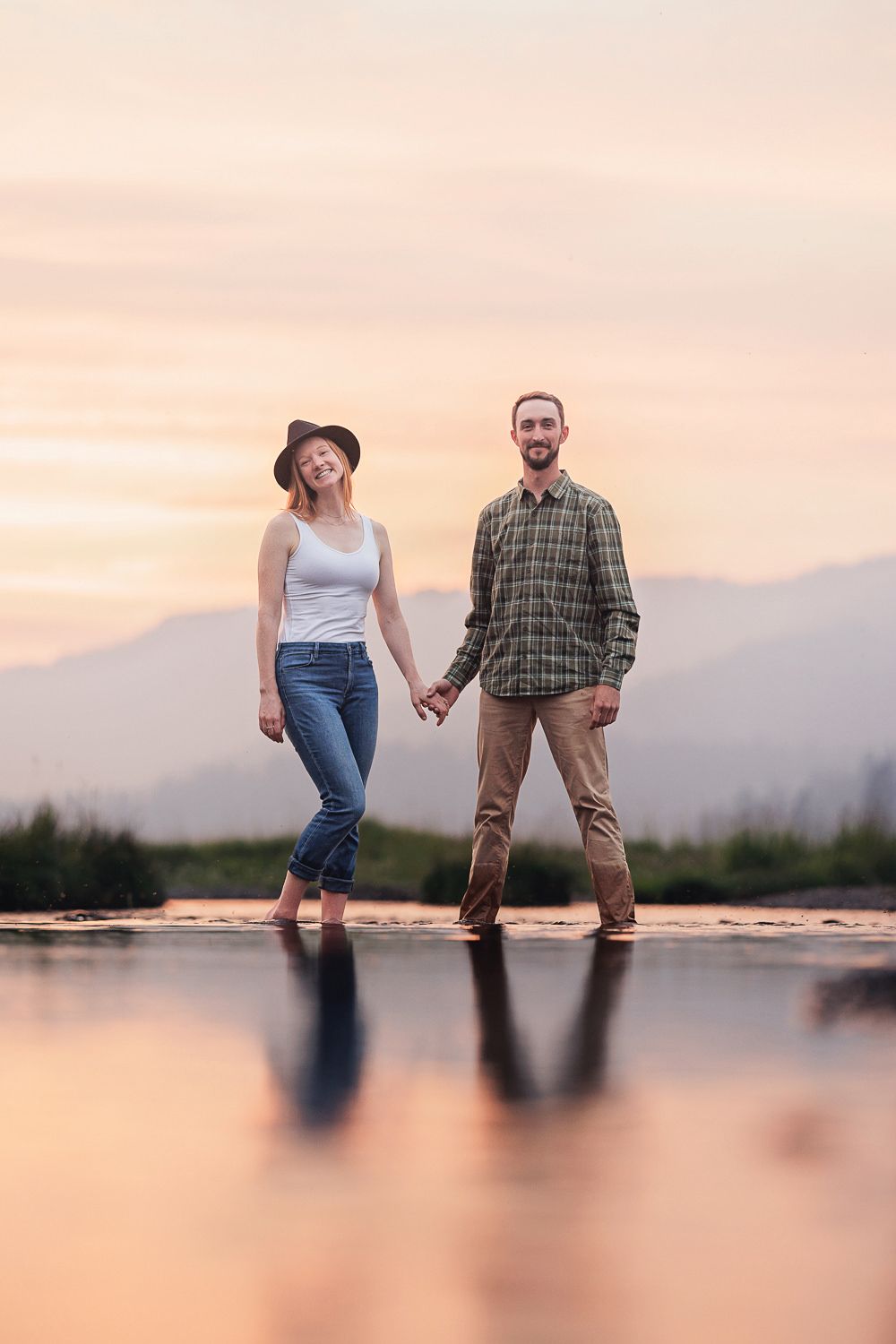 A romantic couple walks along a dock holding hands at sunset with mountains in the distance.